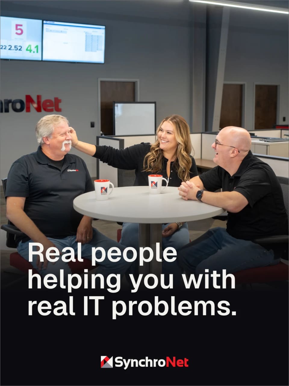 A team photo in the SynchroNet office, three staff laughing around a table with branded mugs, under the line Real people helping you with real IT problems.